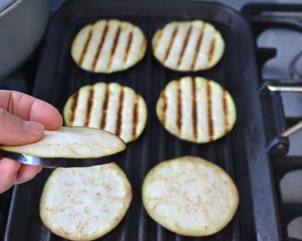 Первым делом баклажан вымыть и тонко нарезать кружочками. Обжарить на хорошо разогретой сковороде-гриль с двух сторон без добавления масла. Сложить на тарелку и ненадолго отставить.