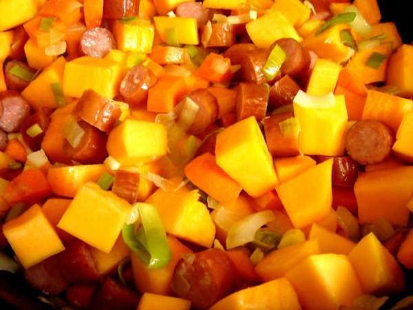 Все овощи нарезать кубиками. В глубоком сотейнике обжарить на растительном масле два вида лука и чеснок,затем-кусочки моркови и после тыквы. Готовить до полуготовности тыквы. Добавить нарезанные на кусочки охотничьи колбаски и курицу, а затем немного растолчённые в ступке специи; тушить 5-7 минут.