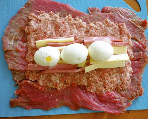 На подготовленную мясную мякоть выложить ровным слоем фарш. Разровнять, оставляя по краям примерно 2 см без начинки, чтобы при скручивании рулета начинка не вышла из него. Сверху и посередине выложить все остальные компоненты начинки; мортаделлу, сыр и сало (если Вы его используете!) В самом конце сверху положить в ряд вареные яйца!  